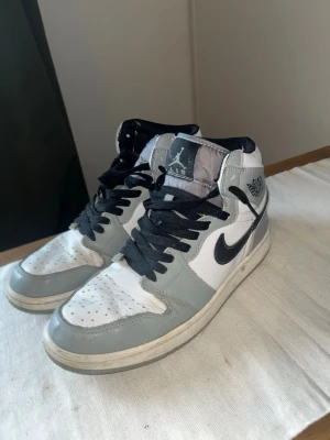 Jordan 1 Mid Light Smoke Grey - Säljer ett par Nike Air Jordan 1 Mid Light Smoke Grey sneakers i vitt och ljusgrått med svarta detaljer och svart swoosh. Skorna har klassisk hög siluett, svart snörning och ikoniska Jordan-loggor på plösen och sidan. Ovandelen är i läder och textil för en clean och sportig look. Skorna är använda som man ser på bilderna men har ingen defekt. Tror jag använde de som skateskor ett tag.