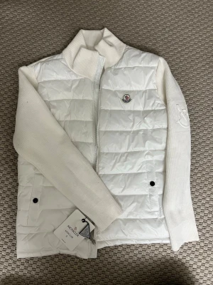 Vit Moncler hybrid cardigan  - Snygg vit dunväst från Moncler med quiltad front och stickade ärmar. Västen har hög krage, dragkedja framtill och två fickor med knapp. Klassisk Moncler-logga på bröstet och diskret emblem på ärmen. Perfekt för lager-på-lager och riktigt clean look. (För endast 1199kr!)