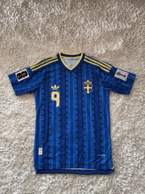 Blå Sverige fotbollströja Adidas - Retro blå fotbollströja från Adidas med Sveriges landslagsemblem och nummer 9 i gult. Tröjan har gula detaljer, tre ränder på axlarna och FIFA-märke på ärmen. Tillverkad i lätt och ventilerande syntetmaterial, perfekt för match eller träning.