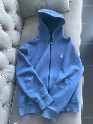 Äkta Marinblå hoodie från Polo Ralph Lauren  - Snygg marinblå hoodie från Polo Ralph Lauren med dragkedja och klassisk broderad logga på bröstet. Tröjan har huva med snörning och två stora fickor framtill. Perfekt för en chill och stilren look. Nypris runt 2500 kr