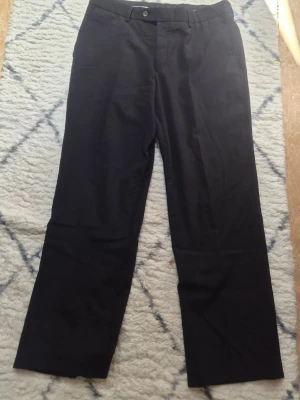 Svarta kostymbyxor från Batiste - Klassiska svarta kostymbyxor från Batiste med rak passform. Tillverkade i en skön mix av 60% ull, 38% polyester och 2% lycra för både stil och komfort. Byxorna har hällor för bälte och stängs med knapp och dragkedja.