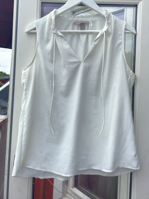 Vit ärmlös blus med knytband - Säljer en stilren vit blus i mjukt material. Blusen är ärmlös, har v-ringning och fina knytband vid halsen samt lätt rynkad kant upptill. Perfekt till jeans eller kjol för en fräsch look.
