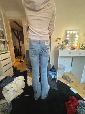 Ljusblå bootcut jeans från True Religion - Säljer ett par ljusblå bootcut jeans från True Religion med klassiska kontrastsömmar och broderade bakfickor. Jeansen har låg midja och är tillverkade i stretchigt denim för en bekväm passform. Perfekta för en avslappnad och trendig look.