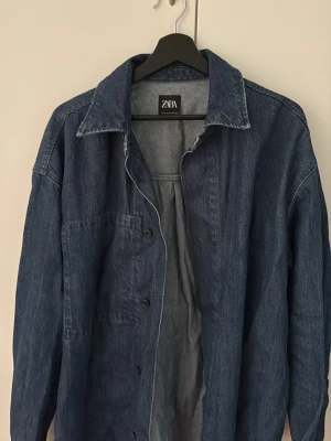 Blå jeansjacka från Zara - Snygg blå jeansjacka från Zara i klassisk modell med krage och knäppning framtill. Jackan har en avslappnad passform och är tillverkad i jeansmaterial. Perfekt att slänga över en hoodie eller t-shirt för en cool look.