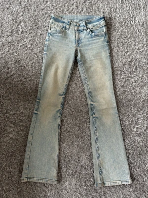 Ljusblå bootcut jeans från H&M - Säljer ett par ljusblå bootcut jeans från H&M med snygga slitningar och klassiska femficksdetaljer. Jeansen har en cool knapp på bakfickan och är tillverkade i denim med en lätt tvättad look. Perfekta för dig som gillar retrostil och trendiga jeans. En liten rosa fläck befinner sig på ena benet av byxan. 