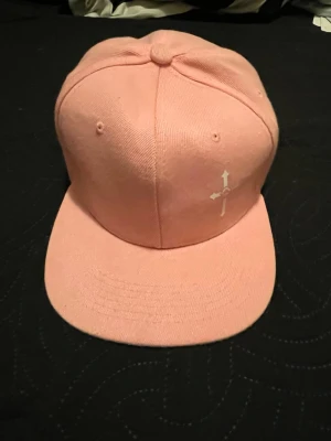 Ljusrosa snapback keps med tryck - Säljer en ljusrosa snapback keps med vit pilgrafik på framsidan. Kepsen har platt skärm och justerbar plastrem bak. Tillverkad i bomullstyg och passar perfekt för en avslappnad streetwear-look.