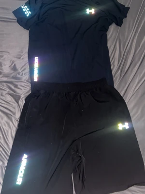Svart träningsset från Under Armour - Snyggt svart träningsset från Under Armour med t-shirt och shorts. Båda plaggen har reflekterande loggor och detaljer i regnbågsfärger. Perfekt för gymmet eller löprundan. Materialet är lätt och andas, vilket gör det bekvämt under träning.
