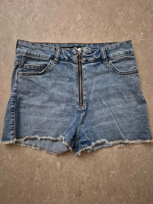 Jeansshorts - Snygga blå denimshorts med hög midja och råa kanter. Shortsens framsida har en cool dragkedja och klassiska fickor både fram och bak. Perfekta för sommarens alla äventyr och ger en avslappnad vibe.