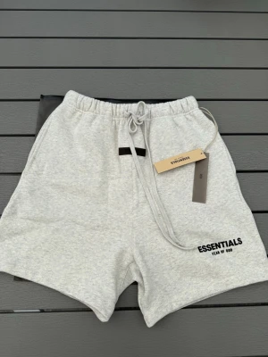Grå Essentials shorts med snörning - Säljer ett par ljusgrå shorts från Fear of God Essentials med svart logga på benet. Shortsen har elastisk midja med snörning och är tillverkade i mjuk bomull, perfekta för en avslappnad stil. Passar till både chill och streetwear.