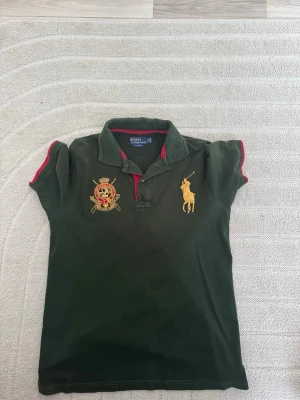 Grön pikétröja från Polo Ralph Lauren - Mörkgrön pikétröja från Polo Ralph Lauren med klassisk krage, korta ärmar och röda detaljer. Stor gul broderad logga på bröstet samt ett broderat emblem på andra sidan. Perfekt för en sportig och stilren look.