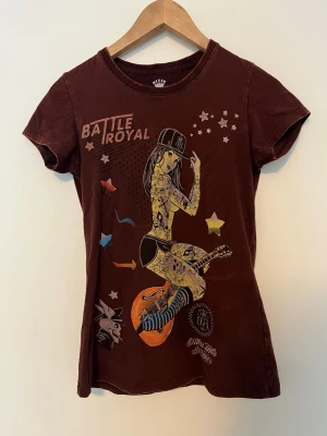 Brun t-shirt med Battle Royal-tryck - Cool brun t-shirt med färgglatt Battle Royal-tryck framtill. Motivet visar en tatuerad tjej med keps och gitarr, omgiven av stjärnor och grafiska detaljer. T-shirten har rund hals och korta ärmar, perfekt för en avslappnad streetwear-look.
