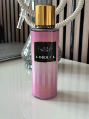 Victoria's Secret Bombshell parfym - Victoria's Secret Bombshell parfym med rosa randig flaska och guldfärgat lock. Flaskan har en elegant och feminin design med svart etikett och guldtext. Perfekt för dig som gillar fräscha och blommiga dofter.