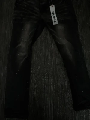 Svarta jeans från Purple med slitningar - Säljer ett par svarta jeans från Purple med coola slitningar och blekta detaljer på låren. Jeansen har små vita färgstänk som ger en trendig look. Perfekta för dig som gillar streetstyle och vill sticka ut.