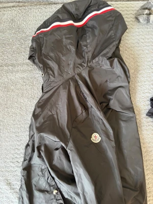 Svart Moncler vindjacka med huva - Snygg svart vindjacka från Moncler med huva och klassisk Moncler-logga på ärmen. Jackan har vita och röda ränder över axlarna och är tillverkad i lätt, glansigt material som skyddar mot vind. Perfekt för dig som vill ha en stilren och sportig look.