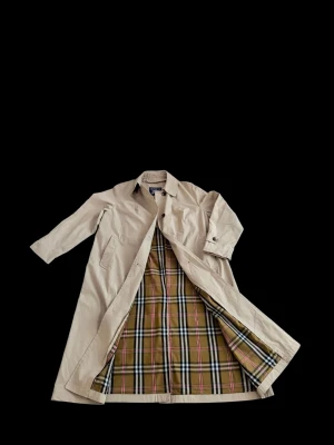 Beige trenchcoat från Burberry - Klassisk beige trenchcoat från Burberry med ikoniskt rutigt foder i beige, svart, vitt och rött. Lång modell med knäppning framtill och två sidofickor. Perfekt för dig som vill ha en tidlös och stilren jacka med exklusiv känsla.