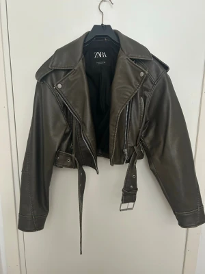 Kort svart skinnjacka från Zara - Snygg kort svart skinnjacka från Zara med bred krage, dragkedja och bälte i midjan. Jackan har silverfärgade detaljer och klassisk bikerstil. Perfekt för dig som vill ha en edgy look till din outfit.