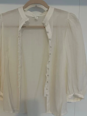 Vit transparent blus med puffärm - Stilren vit blus i transparent material med puffiga långärmar och knappar längs hela framsidan. Klassisk krage och lätt oversized passform ger en trendig look. Perfekt att styla lager på lager för en cool outfit.