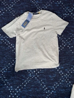 Grå t-shirt från Polo Ralph Lauren - Snygg ljusgrå t-shirt från Polo Ralph Lauren med klassisk logga broderad på bröstet. Tillverkad i mjuk bomull och har en normal passform med korta ärmar och rund hals. Perfekt basplagg för en clean och stilren look.