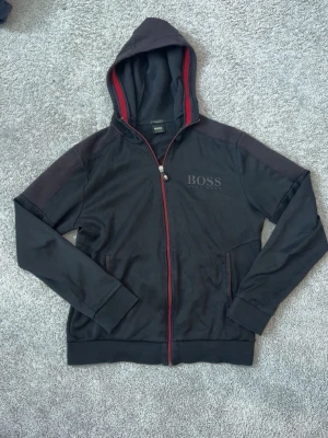 Svart hoodie med dragkedja från BOSS - Snygg svart hoodie från BOSS med dragkedja framtill och röda detaljer längs huvan och dragkedjan. Märket syns diskret på bröstet. Hoodien har två fickor och är tillverkad i mjukt material som känns skönt mot huden. Perfekt för en avslappnad och stilren look. STORLEK M PASSAR ÄVRN S. Ny pris: 1200kr. VÅRT PRIS: 550🔥