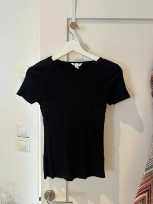 Svart ribbad t-shirt från H&M - En enkel och stilren svart t-shirt från H&M med ribbad struktur. T-shirten har korta ärmar och en rund halsringning. 