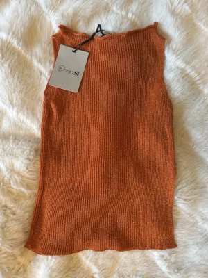 Glittrig ribbad topp från Design by Si - Snygg glittrig ribbad topp i orange från Design by Si. Toppen är ärmlös och har en croppad passform med lätt vågig kant upptill och nedtill. 