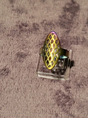 Regnbågsskimrande ring med bikakemönster - Cool ring i metall med regnbågsskimrande yta och ett utstansat bikakemönster. Ringen har en oval, upphöjd front och bred ringband. Perfekt för dig som gillar unika accessoarer med futuristisk känsla.