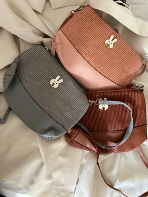 Axelväskor med kaninmärke, passar till accessoarer och barn ungdomar - Säljer tre axelväskor i olika färger: grå, rosa och brun. Väskorna har ribbad textur på framsidan och ett gulligt kaninmärke i metall. De har justerbara axelband och dragkedja. Perfekt för dig som gillar söta detaljer och stilren design. Priset är per styck