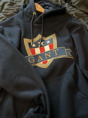 Svart hoodie från GANT med emblem - Snygg svart hoodie från GANT med stort broderat emblem på bröstet i guld, rött, vitt och blått. Klassisk huva med dragsko och känguruficka framtill. Perfekt för dig som gillar streetwear och vill ha en hoodie med premiumkänsla och tydlig logga.