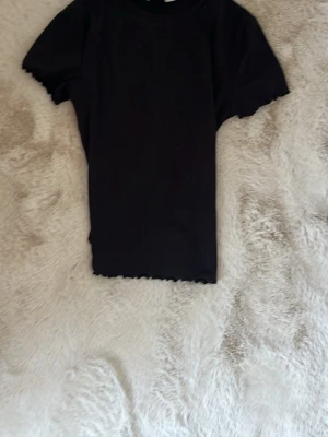 Svart ribbad topp med vågig kant - En svart ribbad topp med korta ärmar och vågiga kanter vid ärmslut och nederkant. Enkel och stilren design som passar till det mesta. Mjuk och stretchig känsla. knappt använd och verkligen så mjuk! passar till typ allt❤️