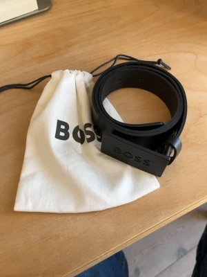 Svart skärp från BOSS med logga - Stilrent svart skärp från BOSS med matt svart spänne där BOSS-loggan är ingraverad. Skärpet är i klassisk design och levereras med en vit tygpåse med BOSS-tryck. Perfekt accessoar för att lyfta din outfit.