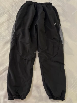 Svarta täckbyxor från Fred Perry för en pojke som är 146cm lång  - Säljer ett par svarta täckbyxor från Fred Perry med elastisk midja och benslut. Byxorna har vita rutiga meshpaneler på sidorna och en liten vit Fred Perry-logga på vänster lår. Materialet är syntet och byxorna har en loose passform. Byxor längd 86 cm 