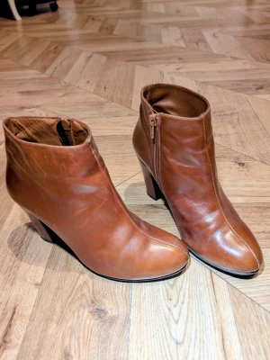 Bruna skinnstövletter med klack - Snygga bruna stövletter i äkta skinn med dragkedja på sidan och smal klack. Klassisk design med rundad tå och stilren söm framtill. Perfekta för dig som vill ha en tidlös look med lite höjd.