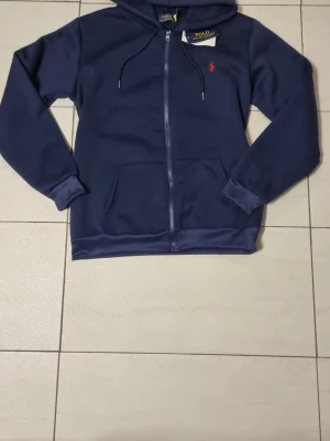 Mörkblå hoodie från Polo Ralph Lauren - Snygg mörkblå hoodie från Polo Ralph Lauren med dragkedja framtill och klassisk röd logga broderad på bröstet. Hoodien har huva med dragsko och två fickor framtill. Perfekt för en avslappnad och stilren look.