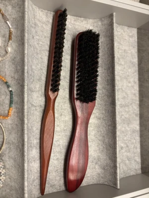 Två borstar i trä - Säljer två hårborstar med handtag i mörkt trä och borst i svart naturfiber. Ena borsten är smal och avlång, perfekt för detaljerad styling, medan den andra är bredare och passar för att borsta större partier hår. Snygg klassisk look och sköna att hålla i.