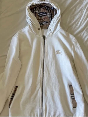 Vit hoodie från Burberry med dragkedja - Säljer en vit hoodie från Burberry med klassiskt rutigt foder i huvan och detaljer vid fickorna. Hoodien har dragkedja framtill, långa ärmar och en liten Burberry-logga broderad på bröstet. Perfekt för en clean och stilren look.