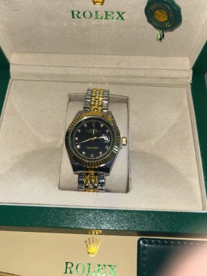 Elegant Role Damklocka i  Stål – Komplett Set med Box och Certifikat - Snygg role Datejust med svart urtavla och tvåtonad länk i guld och silver. Klockan har klassisk räfflad bezel och datumvisning. Levereras i originalbox med certifikat och tillbehör. Perfekt accessoar för dig som gillar exklusiv stil.