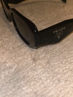 Svarta solglasögon från Prada - Snygga svarta solglasögon från Prada med breda skalmar och diskret logga på sidan. Glasögonen har en modern, kantig form och är tillverkade i blankt syntetmaterial. Perfekta för dig som vill ha en stilren och lyxig accessoar. Priset kan diskuteras vid snabb affär 