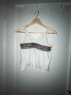 Vit linnetopp med färgglada band - Söt vit linnetopp med smala axelband och omlott framtill. Toppen har dekorativa band i turkos, lila och brunt med paljettdetaljer under bysten. Perfekt för varma dagar och enkel att styla till jeans eller kjol.