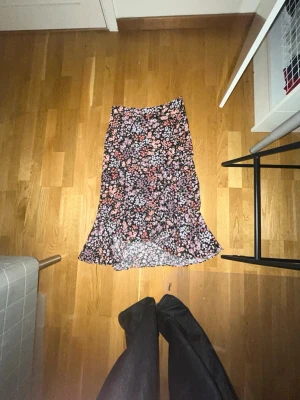 Blommig svart långkjol med volang - Supersnygg långkjol med svart botten och blommigt mönster i rosa, lila och vitt. Kjolen har volang nertill och en smickrande passform. Perfekt för dig som vill ha en feminin och trendig look.