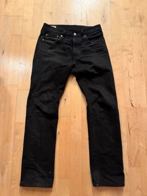 Svarta 501 Levi's jeans  - Säljer ett par svarta jeans från Levi's med klassisk femficksmodell och raka ben. Jeansen har en läderpatch bak i midjan och den ikoniska röda Levi's-taggen på bakfickan. Tillverkade i slitstark denim med knappgylf och silverfärgade nitar.