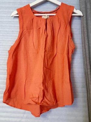 Orange ärmlös blus med veck - Snygg orange blus med rund halsringning och dekorativa veck framtill. Blusen är ärmlös och har en lätt, luftig passform som passar perfekt till varma dagar. Materialet känns mjukt och bekvämt mot huden.