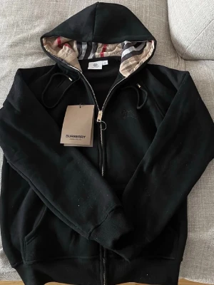 Svart hoodie från Burberry - Snygg svart hoodie från Burberry med dragkedja och klassiskt rutigt foder i huvan. Broderad logga på bröstet och fickor framtill. Tillverkad i mjuk bomull för skön känsla. Perfekt för dig som vill ha en stilren och exklusiv look. Använd sirka 7 eller 10 gånger 