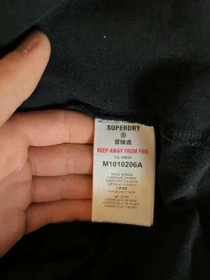 Svart t-shirt från Superdry - En klassisk svart t-shirt från Superdry med diskret broderad logga på bröstet. T-shirten har rund hals och korta ärmar, perfekt för en clean och enkel stil. Tillverkad i mjuk bomull för skön komfort.