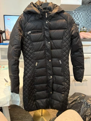 Jaka storlek 36 Rockand Blue - Säljer en svart lång dunjacka med huva och dragkedjor framtill. Jackan har quiltade partier på sidorna och tre fickor med dragkedja. Perfekt för kalla vinterdagar och har en snygg, figurnära passform.