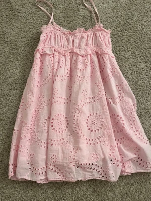 Ljusrosa kort klänning med spets - Söt ljusrosa kort klänning med tunna axelband och broderade spetsdetaljer. Klänningen har ett luftigt och loose passform med volang upptill och ett romantiskt mönster. Perfekt för sommarens varma dagar.