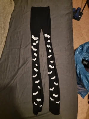 Svarta tights med vita fladdermöss - Säljer ett par svarta tights med vita fladdermusmotiv längs benen. Tightsen har en klassisk passform och är stretchiga, perfekta för dig som vill ha en cool och unik look. Materialet känns mjukt och bekvämt mot huden.