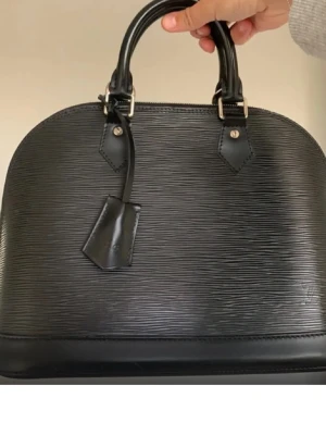 Svart Louis Vuitton Alma handväska - Snygg svart handväska från Louis Vuitton i klassisk Alma-modell. Väskan är tillverkad i strukturerat Epi-läder med rundad form och dubbla handtag. Diskret Louis Vuitton-logga på läderdetaljen framtill. Perfekt för dig som gillar tidlös och lyxig design. Medföljer lås, nyckel och äktighetskort. 