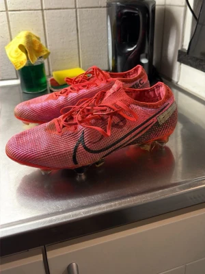 Nike Mercurial röda fotbollsskor - Säljer ett par Nike Mercurial fotbollsskor i rött med svarta detaljer och stora Nike-loggor på sidorna. Skorna har snörning och metallskruvdobbar för bästa grepp på planen. Ovandelen är i ett lätt syntetmaterial som ger bra bollkontroll och speed.