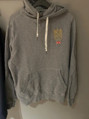 Grå hoodie från Morris med tryck - Snygg grå hoodie från Morris med guldfärgat tryck och brittisk flagga på bröstet. Klassisk känguruficka framtill, vita dragsnören i huvan och ribbade muddar vid ärmar och nederkant. Perfekt för en avslappnad och stilren look.