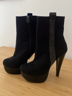 Svarta platåstövletter med strassdetalj - Snygga svarta stövletter med hög platå och smal klack. Ovandelen är i mockaimitation och på sidan finns en rad med svarta strasstenar som ger extra bling. Dragkedja på insidan för enkel påtagning. Perfekta för dig som vill sticka ut.